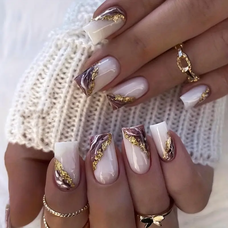 A close-up of manicured nails showcases a glossy finish and intricate design with white and maroon swirls, plus gold accents. The long nails resemble Snazzi Nails Purple Marble Nails. The person wears a textured white garment and several rings.