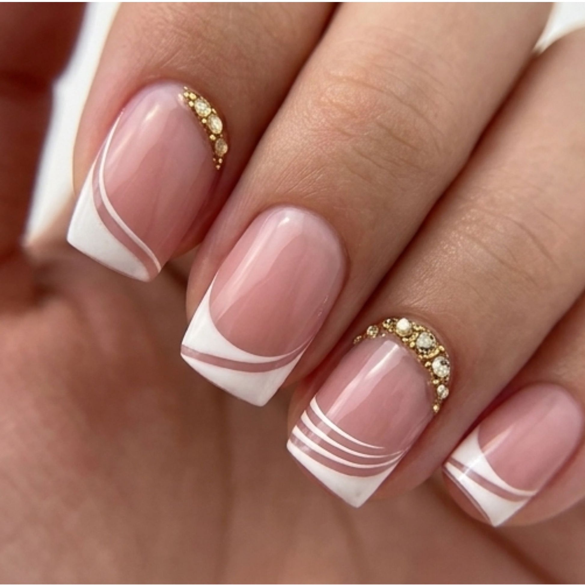 Short square blush pink press-on nails with white French tips, curved wave accents, fine white lines, and gold rhinestone cuticle details.