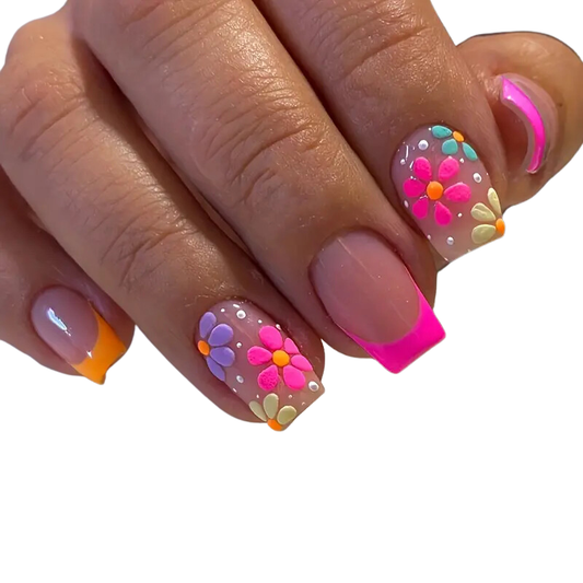A hand displays Snazzi Nails Flowery Day DIY full cover nails with bright florals in pink, orange, purple, and blue alongside white dots. Some short square nails have solid pink and orange tips. The simple white background elevates the vibrant artistry.