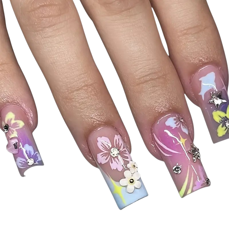 A close-up of a hand showing off long, coffin-shaped press-on nails with pastel floral and abstract designs. The nails feature pink, purple, blue, and yellow patterns, decorated with 3D flowers and rhinestones, set against a plain white background.