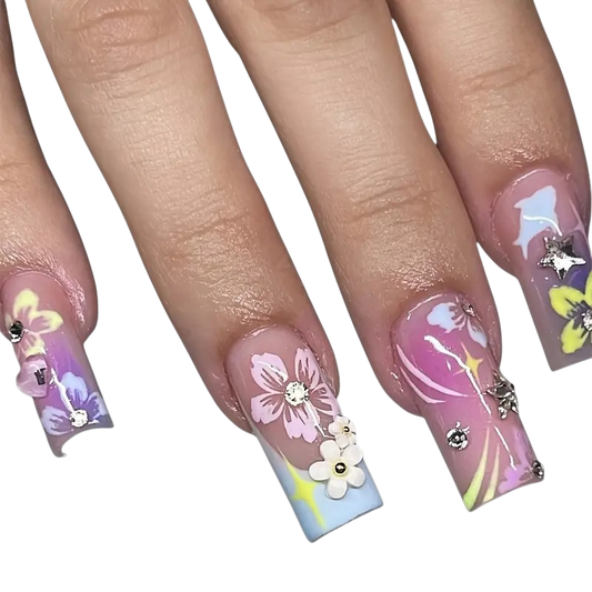 A close-up of a hand showing off long, coffin-shaped press-on nails with pastel floral and abstract designs. The nails feature pink, purple, blue, and yellow patterns, decorated with 3D flowers and rhinestones, set against a plain white background.
