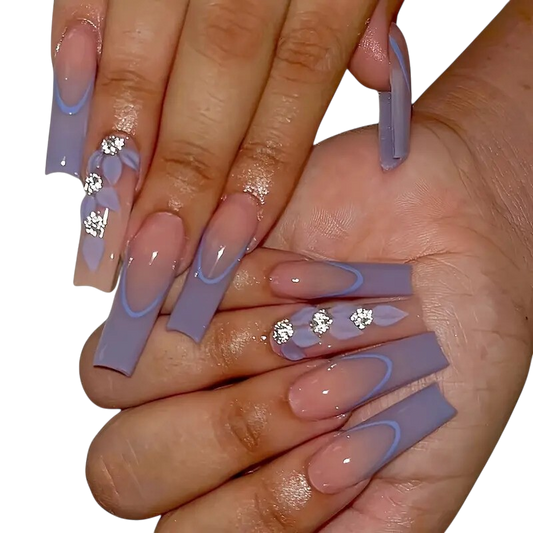 A close-up of a person's hands showcasing long, square-shaped press-on nails with a lavender and nude gradient design, French tips, and rhinestone embellishments, set against a plain white background.
