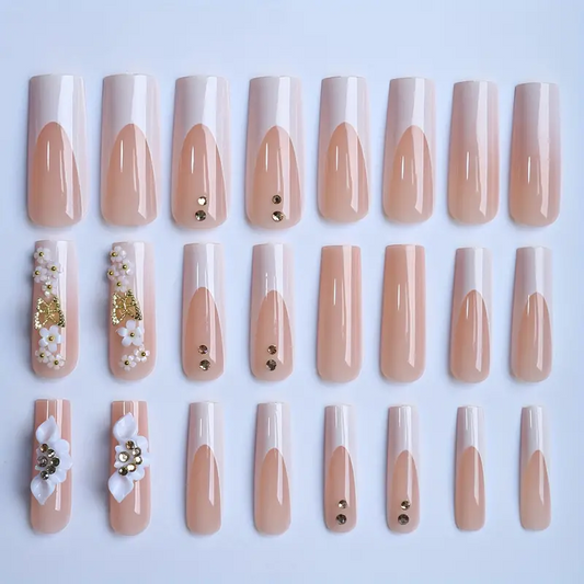 A set of 20 false press-on nails arranged in four rows of five on a light blue background. The nails feature a glossy French tip design with a nude beige and white gradient, with some decorated with small gold rhinestones, white and gold flowers, and white pearls.