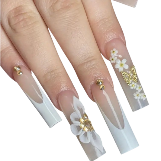 A close-up of a hand showcasing long square press-on nails with a white French tip design, adorned with 3D white flowers, gold glitter accents, and small rhinestone clusters.