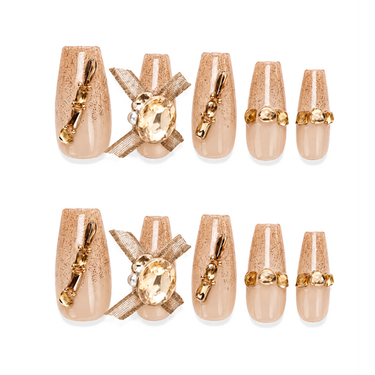 Snazzi Pink Shop Honey Couture long coffin nails with nude glossy base, golden-bronze speckles, gems, and mesh bows.