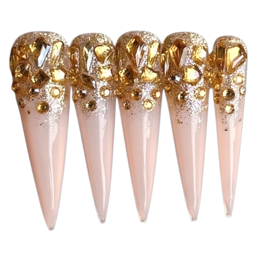 Snazzi Pink Shop Jeweled Reign 2XL stiletto press-on nails with nude base, gold glitter, and amber-gold crystals.